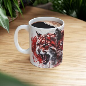 Puede incluir: Taza de café de cerámica blanca con una ilustración colorida de dos caras de lince. El lince de la izquierda está rugiendo con la boca abierta. El fondo es un diseño abstracto rojo y blanco.