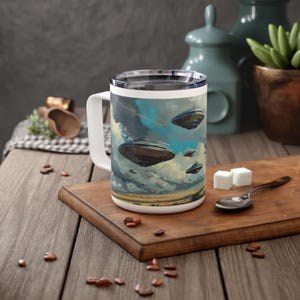 May include: White ceramic mug with a handle, featuring a colourful illustration of multiple flying saucers in a cloudy sky. The mug has a stainless steel lid.