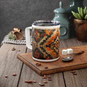 May include: A white ceramic mug with a handle features a colorful geometric pattern inspired by traditional rugs. The mug is sitting on a wooden surface with coffee beans scattered around it.