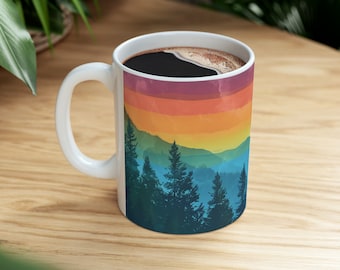 May include: White ceramic coffee mug with a landscape design featuring a colorful sunset over a mountain range and evergreen trees. The mug is filled with dark coffee and has a white handle.
