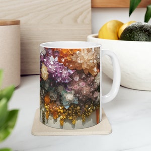 May include: A white ceramic mug with a colorful image of a cluster of crystals. The crystals are in shades of purple, pink, orange, yellow, and blue. The mug has a handle on the right side.