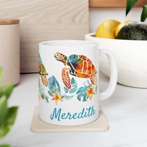 May include: White ceramic mug with a colorful design of two sea turtles swimming among tropical leaves and flowers. The mug has the name "Meredith" printed on the bottom in blue.