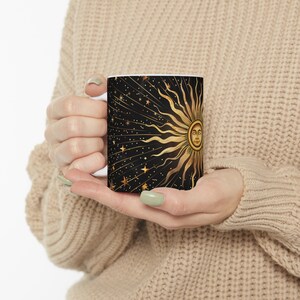 Könnte beinhalten: Eine weiße Keramiktasse mit einem schwarzen und goldenen Design. Das Design zeigt eine stilisierte Sonne mit einem Gesicht und Lichtstrahlen, umgeben von Sternen auf schwarzem Hintergrund.