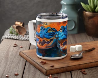 Puede incluir: Una taza de cerámica blanca con un diseño azul y naranja que presenta dos caimanes con la boca abierta. La taza tiene una tapa y un asa.