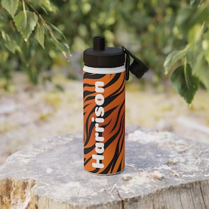 Op de afbeelding: Een waterfles met een zwarte dop en een oranje en zwarte tijgerstreep. De naam "Harrison" staat verticaal in het wit gedrukt. De fles staat op een boomstronk.
