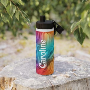 Op de afbeelding: Een waterfles met een zwarte dop en een kleurrijk, wervelend ontwerp in tinten rood, oranje, geel, blauw en groen. De naam "Caroline" staat verticaal in het wit op de fles.