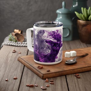 May include: White ceramic mug with a handle. The mug features a purple and gray abstract design of a person with long hair, holding a bow and arrow.