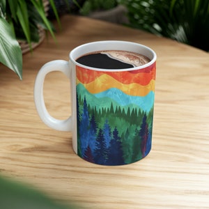 May include: White ceramic mug with a rainbow mountain and forest design. The mug is filled with black coffee.