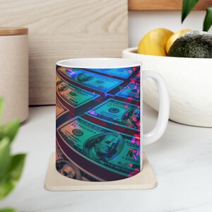 May include: White ceramic mug with a colorful design of US dollar bills. The bills are arranged in a stack and have a blue, green, and red color scheme.