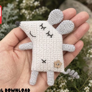 Puede incluir: Un pequeño adorno de ratón de ganchillo, plano, hecho con hilo gris claro y blanco roto. El ratón presenta ojos cerrados bordados, una sonrisa cosida, una nariz gris oscuro, dos brazos pequeños, dos piernas, una cola rizada y un pequeño botón de flor de madera.