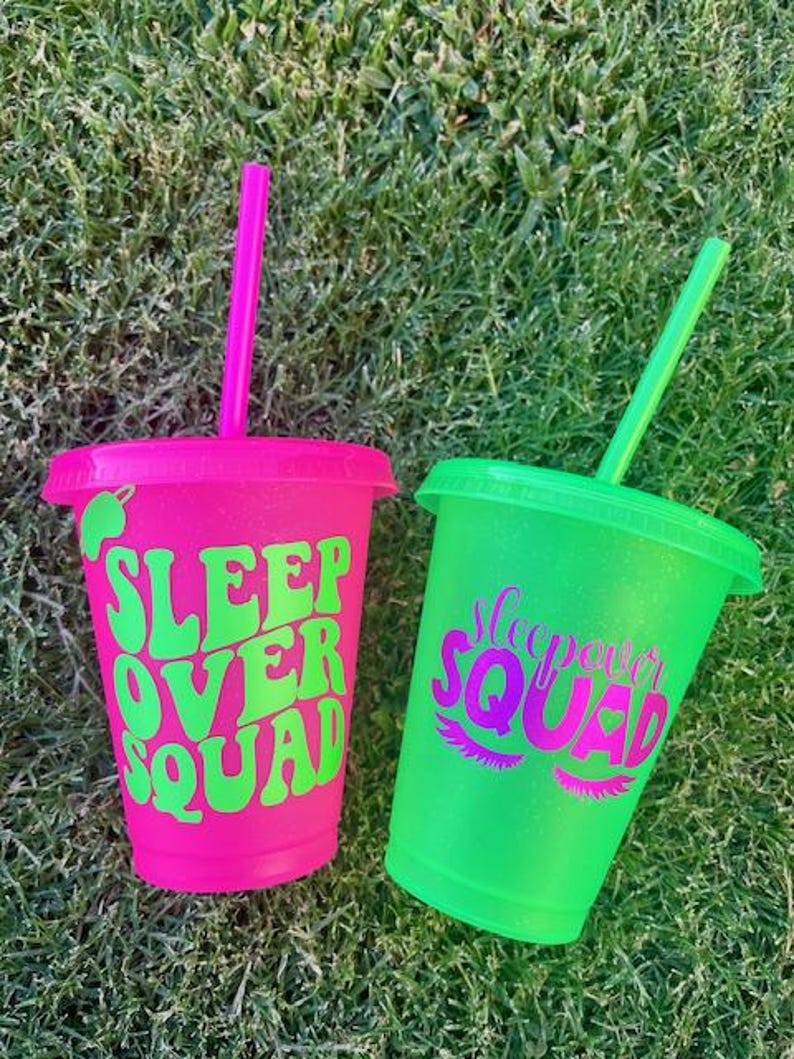 K&ouml;nnte beinhalten: Zwei glitzernde Plastikbecher mit Strohhalmen. Einer ist leuchtend rosa mit den Worten "SLEEP OVER SQUAD" in Gr&uuml;n. Der andere ist gr&uuml;n mit den Worten "Sleepover SQUAD" in Pink. Beide Becher stehen auf gr&uuml;nem Gras.