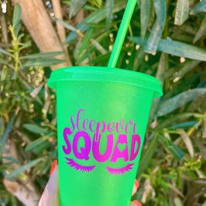 K&ouml;nnte beinhalten: Ein neongr&uuml;ner Kunststoffbecher mit passendem Deckel und Strohhalm. Der Becher tr&auml;gt den Text "Sleepover Squad" in Pink mit Wimpern und einem Herz. Der Becher wird von einer Person gehalten.
