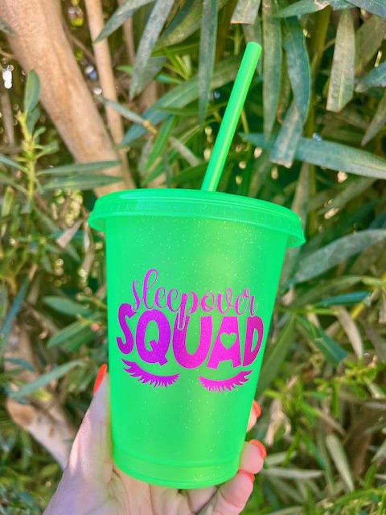 K&ouml;nnte beinhalten: Ein neongr&uuml;ner Kunststoffbecher mit passendem Deckel und Strohhalm. Der Becher tr&auml;gt den Text "Sleepover Squad" in Pink mit Wimpern und einem Herz. Der Becher wird von einer Person gehalten.