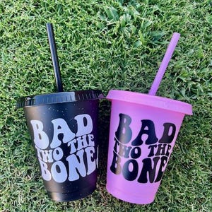 K&ouml;nnte beinhalten: Zwei Kunststoffbecher mit Deckeln und Strohhalmen. Ein Becher ist mattschwarz mit wei&szlig;em Text "BAD TO THE BONE" und einem schwarzen Strohhalm. Der andere Becher ist matt hellviolett mit schwarzem Text "BAD TO THE BONE" und einem violetten Strohhalm.