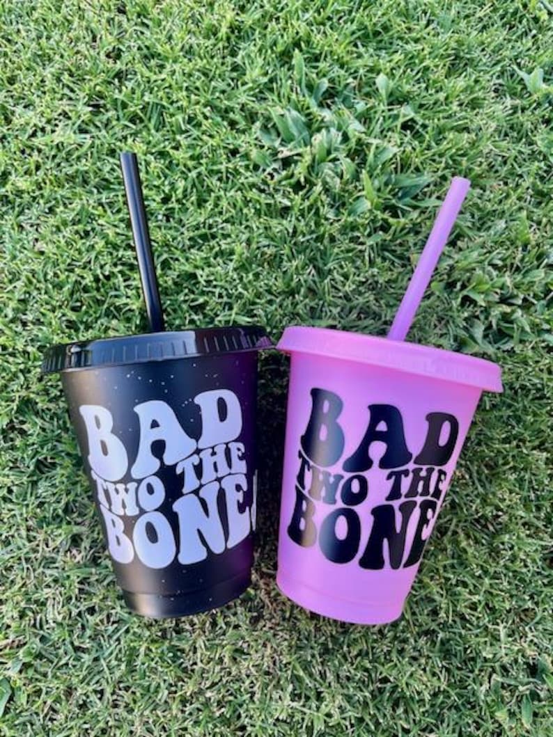 K&ouml;nnte beinhalten: Zwei Kunststoffbecher mit Deckeln und Strohhalmen. Ein Becher ist mattschwarz mit wei&szlig;em Text "BAD TO THE BONE" und einem schwarzen Strohhalm. Der andere Becher ist matt hellviolett mit schwarzem Text "BAD TO THE BONE" und einem violetten Strohhalm.
