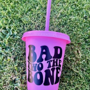 K&ouml;nnte beinhalten: Ein leuchtend pinkfarbener, wiederverwendbarer Kunststoffbecher mit passendem Deckel und Trinkhalm. Auf der Vorderseite ist der Text "BAD TWO THE BONE" in fetten, schwarzen Retro-Buchstaben aufgedruckt, was ein kontrastreiches Design ergibt.