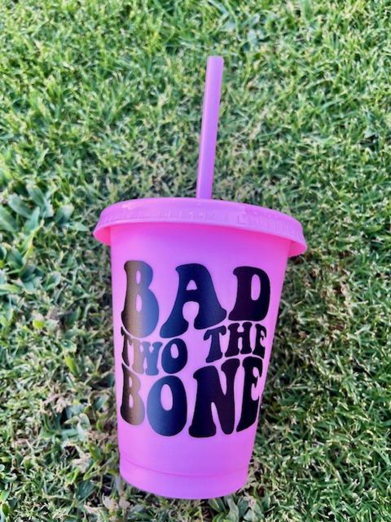K&ouml;nnte beinhalten: Ein leuchtend pinkfarbener, wiederverwendbarer Kunststoffbecher mit passendem Deckel und Trinkhalm. Auf der Vorderseite ist der Text "BAD TWO THE BONE" in fetten, schwarzen Retro-Buchstaben aufgedruckt, was ein kontrastreiches Design ergibt.