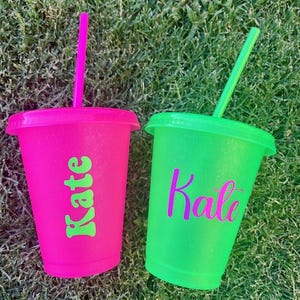 K&ouml;nnte beinhalten: Zwei glitzernde Plastikbecher mit Deckel und Strohhalm. Einer ist pink mit dem Namen "Kate" in Gr&uuml;n, der andere neongr&uuml;n mit "Kate" in Lila. Beide stehen auf gr&uuml;nem Gras.