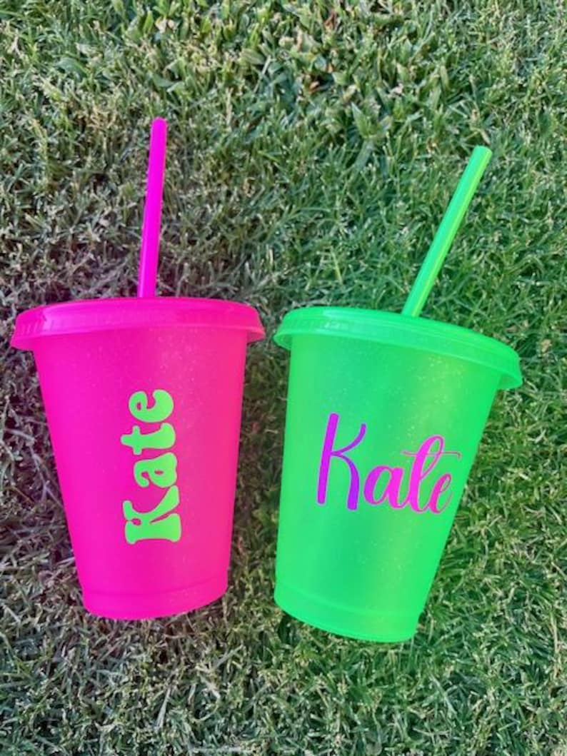 K&ouml;nnte beinhalten: Zwei glitzernde Plastikbecher mit Deckel und Strohhalm. Einer ist pink mit dem Namen "Kate" in Gr&uuml;n, der andere neongr&uuml;n mit "Kate" in Lila. Beide stehen auf gr&uuml;nem Gras.