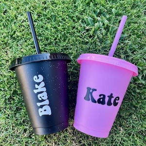 K&ouml;nnte beinhalten: Zwei Kunststoffbecher mit Deckeln und Strohhalmen. Ein Becher ist schwarz mit einem dezenten Sprenkelmuster und dem wei&szlig;en Text Blake. Der andere Becher ist einfarbig hellviolett mit dem schwarzen Text Kate. Beide Becher haben einen passenden Strohhalm.