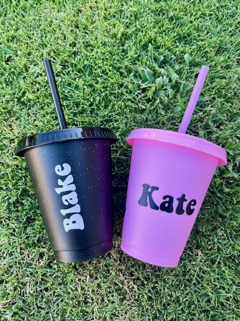 K&ouml;nnte beinhalten: Zwei Kunststoffbecher mit Deckeln und Strohhalmen. Ein Becher ist schwarz mit einem dezenten Sprenkelmuster und dem wei&szlig;en Text Blake. Der andere Becher ist einfarbig hellviolett mit dem schwarzen Text Kate. Beide Becher haben einen passenden Strohhalm.