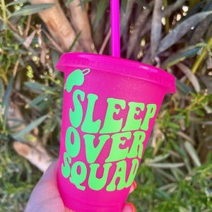 K&ouml;nnte beinhalten: Ein pinkfarbener Kunststoffbecher mit passendem Deckel und Strohhalm. Der Becher tr&auml;gt den Text "SLEEP OVER SQUAD" in Gr&uuml;n, mit einer gr&uuml;nen Chili-Pfeffer-Grafik. Der Becher wird vor einem nat&uuml;rlichen Hintergrund gehalten.