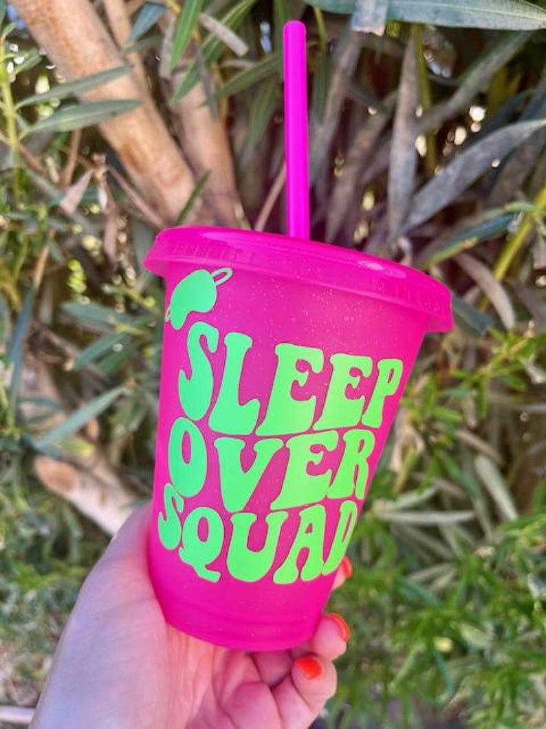 K&ouml;nnte beinhalten: Ein pinkfarbener Kunststoffbecher mit passendem Deckel und Strohhalm. Der Becher tr&auml;gt den Text "SLEEP OVER SQUAD" in Gr&uuml;n, mit einer gr&uuml;nen Chili-Pfeffer-Grafik. Der Becher wird vor einem nat&uuml;rlichen Hintergrund gehalten.