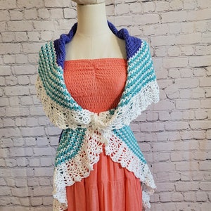 May include: A white, teal, and purple crochet shawl with a scalloped edge. The shawl is draped over a coral-colored dress.