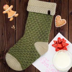 May include: A green and white crocheted Christmas stocking with a white cuff and a brown leather loop. The stocking is hanging on a wooden surface.