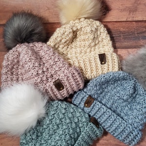 May include: A collection of handmade crochet beanies in various colors, including pink, blue, cream, and teal. Each beanie features a textured stitch pattern and a faux fur pom-pom. Small brown tags are attached to the beanies.