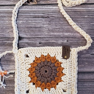 Puede incluir: Bolso bandolera de crochet color crema con un diseño de girasol en tonos marrón y naranja. El bolso tiene una correa trenzada y una pequeña etiqueta de cuero. El bolso es de forma cuadrada y tiene un aspecto texturizado.