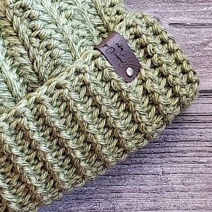 Może przedstawiać: Zielony wełniany kapelusz z brązową skórzaną etykietą z napisem "Sewn with Love".