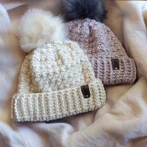 Peut inclure: Deux bonnets en crochet avec des pompons en fausse fourrure. Un bonnet est beige et l'autre est violet clair. Les deux bonnets ont un patch en cuir avec un logo sur le côté.