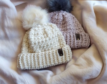May include: Two crocheted hats with faux fur pom poms. One hat is beige and the other is light purple. Both hats have a leather patch with a logo on the side.