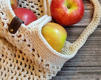 May include: A beige crocheted mesh bag with a brown leather tag that says "Summer Oak Designs" is filled with red and yellow apples. The bag is sitting on a wooden surface.
