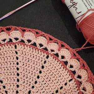 May include: A close-up of a crochet project in progress, featuring a circular design in shades of pink and rose. A ball of pink yarn and a crochet hook are visible, suggesting a handmade craft project. The yarn label reads "10% RAYON FROM BAMBOO".