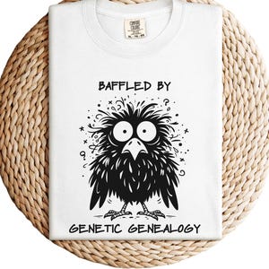 May include: White t-shirt with a black and white graphic of a confused bird and the text "BAFFLED BY GENETIC GENEALOGY". The shirt is folded on a woven, circular surface.