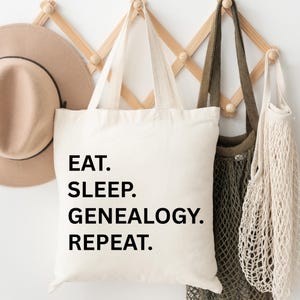 Könnte beinhalten: Eine cremefarbene Tragetasche mit den Worten "EAT. SLEEP. GENEALOGY. REPEAT." in Schwarz gedruckt. Die Tasche hängt an einem Holzgestell neben anderen Taschen und einem beigen Hut.