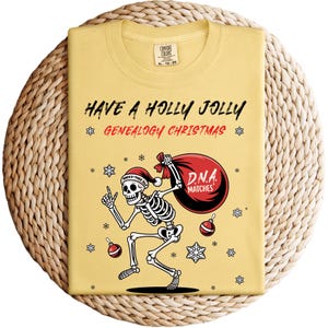 以下が含まれることがあります： 「HAVE A HOLLY JOLLY GENEALOGY CHRISTMAS」の文字と、サンタ帽をかぶった骸骨が「D.N.A. MATCHES」と書かれたバッグを持っている黄色のTシャツ。デザインには雪の結晶とオーナメントが含まれています。