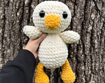 Puede incluir: Juguete de peluche de pato blanco tejido a crochet con patas amarillas y un pico amarillo. El pato tiene ojos negros y está sentado en un tronco de árbol.
