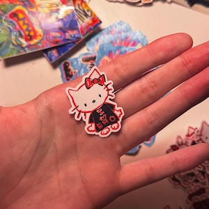 May include: A white Hello Kitty sticker with a red bow and black and red clothing. The sticker is on a hand with a black sleeve.
