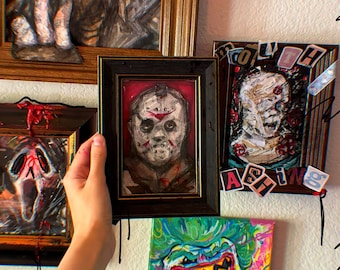 May include: A collection of framed horror movie character paintings. The paintings feature Jason Voorhees, Ghostface, and a zombie-like figure. The paintings are displayed on a white wall with black and white doodles.