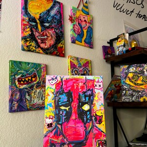 May include: A collection of colorful, abstract paintings on canvas. The paintings feature various characters and designs, including a pink and black Deadpool, a green and yellow monster, a yellow and red Wolverine, and a yellow and red character with a party hat. The text 'CANVASES SALE velvet toad ART' is written in black marker on a white background.