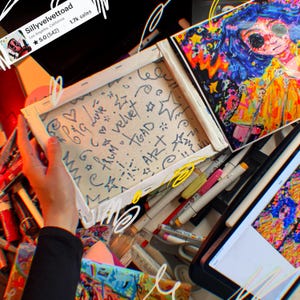 May include: An overhead shot of an art studio with a canvas featuring handwritten text and doodles. The canvas is held by a person. Other art supplies, including markers and a digital tablet, are scattered around. A colorful artwork is also visible.