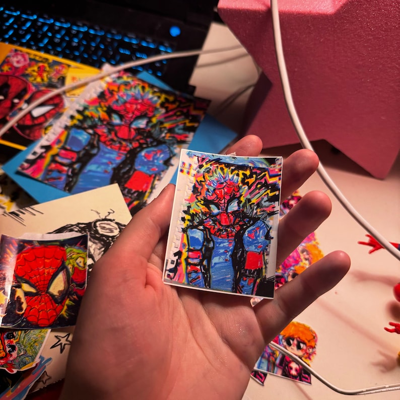 May include: A colourful sticker featuring a cartoon illustration of Spider-Man with a red and blue suit, surrounded by a bright, abstract design. The sticker is on a white background.