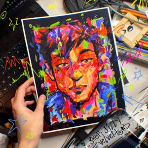 Könnte beinhalten: Ein farbenfrohes Porträtgemälde auf schwarzem Papier. Das Kunstwerk zeigt ein Gesicht mit lebendigen Pinselstrichen in Rot-, Orange-, Blau- und Grüntönen. Das Gemälde wird vor einem Hintergrund mit einer Tastatur und Kunstzubehör gehalten.