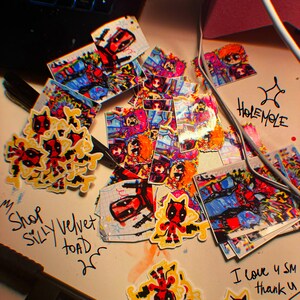 May include: A collection of colorful stickers featuring cartoon characters, including Deadpool and a cat. The stickers are on a white surface with handwritten text that says "Shop Silly Velvet Toad".