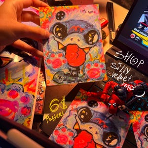K&ouml;nnte beinhalten: Sechs bunte Poster mit einer Comicfigur mit einer gro&szlig;en Zunge und leuchtenden Augen. Die Figur ist von einem bunten Hintergrund mit einem psychedelischen Muster umgeben. Die Poster sind in einem Shop namens "Silly Velvet Toad" erh&auml;ltlich.