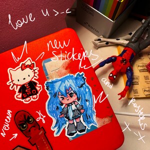 May include: A red notebook with a variety of stickers and drawings on it. The stickers include a cartoon girl with blue hair, a cartoon cat with a red bow, and a Spiderman character. The notebook has the words "love u>-<" and "new stickers" written on it.