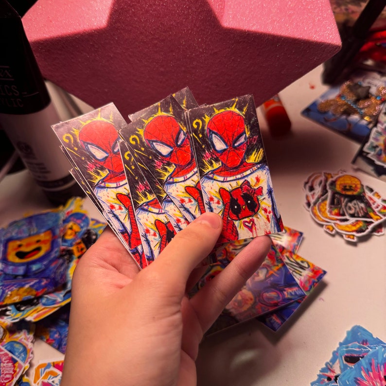 Puede incluir: Una pila de pegatinas coloridas con un personaje de dibujos animados de Spiderman con un signo de interrogaci&oacute;n sobre su cabeza. El personaje de Spiderman lleva una camisa blanca con salpicaduras de pintura roja y negra. Las pegatinas est&aacute;n impresas en papel blanco.